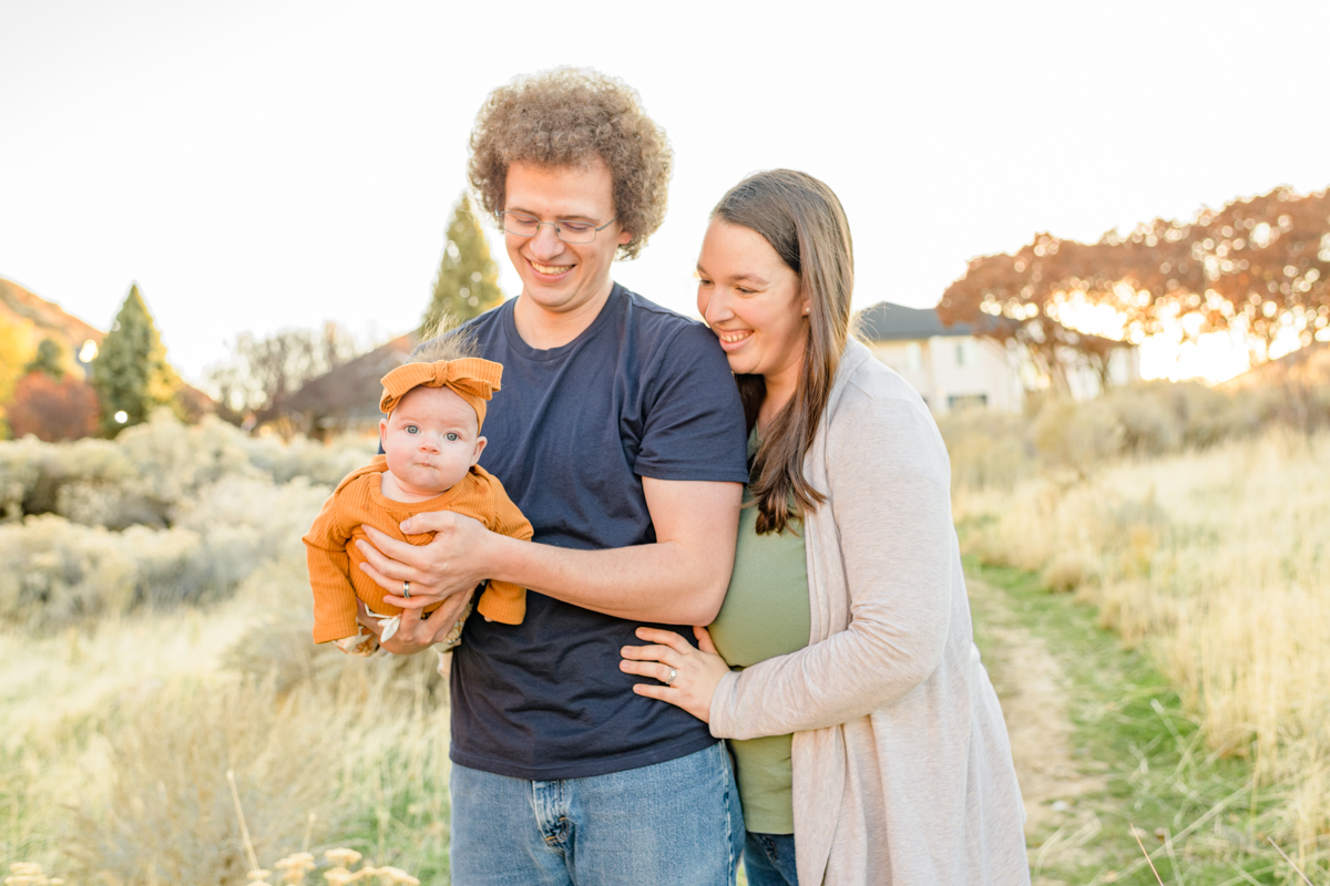 Family photo of a young father holding his infant daughter with his wife looking over his shoulder smiling during a full session with Salt Lake Photographer Jodi Cornwall Photography.