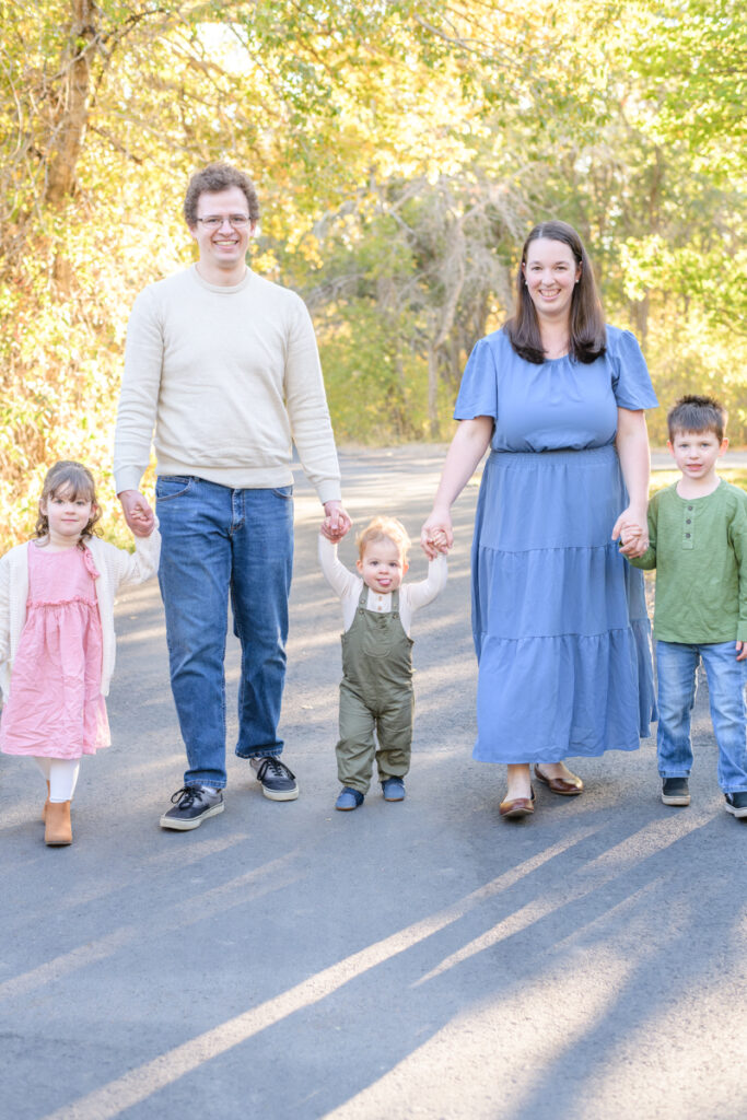 A family with three young children hold hands and walk through a forest during a vibrant fall family session with Jodi Cornwall Photography.