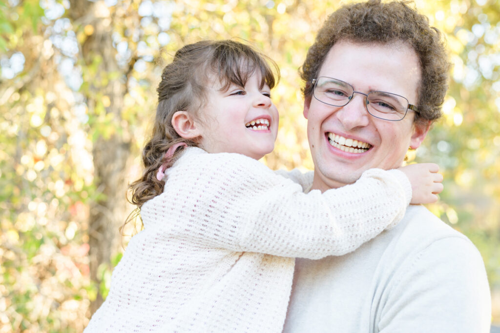 A dad holds his young daughter, both smiling and laughing during a vibrant fall family session with Jodi Cornwall Photography.