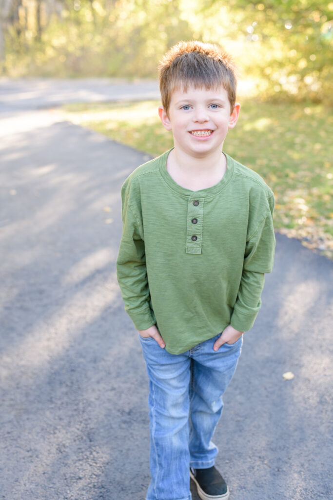 A young boy smiles on a pathway in a forest during a vibrant fall family session with Jodi Cornwall Photography.